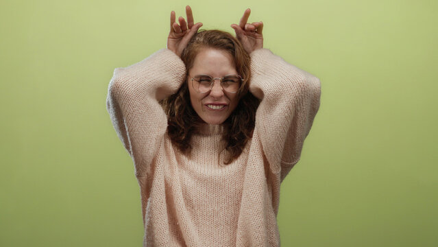 Young woman making playful gestures with hands on head, wearing glasses and sweater, standing against isolated yellow background, exuding a fun and cheerful mood.