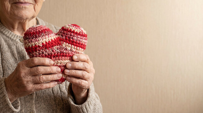 An older person holds a red and pink handmade heart in hands. This moment takes place on Valentine&rsquo;s Day, a time for love and connection. The background is simple and warm