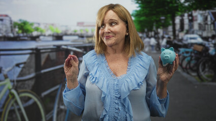 Middle age woman holding a blue piggy bank in her right hand and raising left palm in a pledge gesture on a street by a canal; financial confidence.