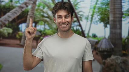 Man points index finger upward while smiling, wearing white t shirt in forest with palm trees and...