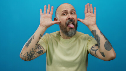 Man posing playfully with tattoos and beard against bright blue background, showing expressive face and playful gestures in a humorous setting.