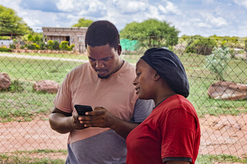 village african woman and social worker men, using a smartphone , outdoors in the nature, at home ,wire fence behind