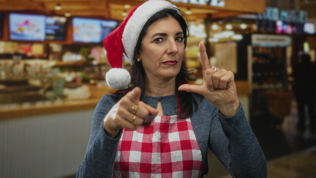 Woman wearing red apron and santa hat makes loser gesture with her hand and points index finger toward viewer in restaurant; mockery. - Powered by Adobe