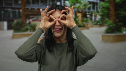 Woman in green cutout sleeve top framing eyes with hands in hand binoculars gesture on street;...