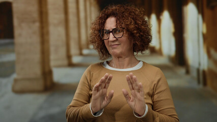 Hispanic middle age woman in an old university setting makes a rejection gesture outdoors, capturing emotion and heritage against ancient architecture backdrop, wearing glasses.