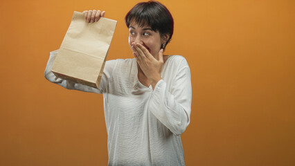 Woman holding paper bag aloft with hand covering mouth, smiling and surprised in a studio orange backdrop mid reveal; surprise.
