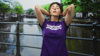 Woman in purple volunteer tshirt with short hair, hands behind head and closed eyes, leaning by a railing on a street beside a canal and bridge; calm relief.