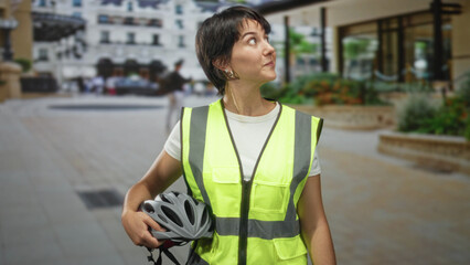 Woman with short hair in a neon reflective vest holds a bicycle helmet, smiles and looks up in a street plaza; urban commuting confidence.