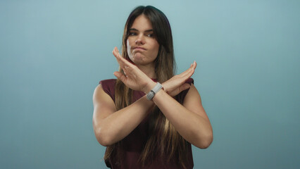 Hispanic woman crossing arms to form an x in rejection over isolated blue background, expressing refusal or disagreement with clear body language.