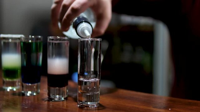Skilled barman preparing layered cocktail shots at a bar at night, pouring colorful liquor to create beautiful gradient drinks under warm light. High quality 4k footage