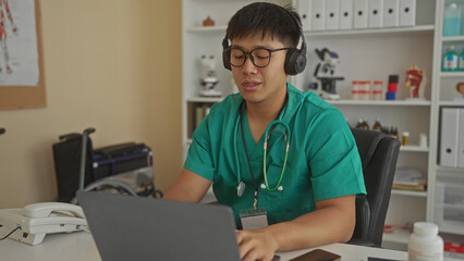 Obraz premium Young man in a clinic wearing scrubs and headphones using a laptop in a workplace setting with medical equipment around.