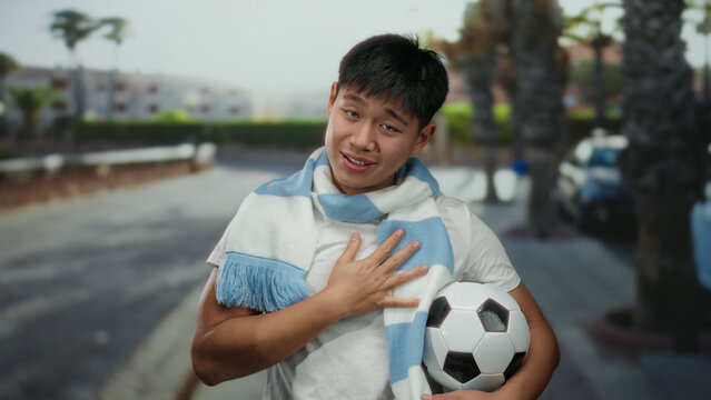 Young man holding soccer ball on city street, wearing a blue scarf, exhibits outdoor fan enthusiasm, showcasing sports passion and joyful expression.