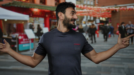 Man with a beard smiling and pointing while standing outdoors on a vibrant city street with bustling people and festive decorations in the background.