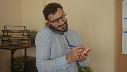 Hispanic man writing notes while multitasking in a modern office setting, holding a phone between his ear and shoulder, creating a busy yet focused work atmosphere.