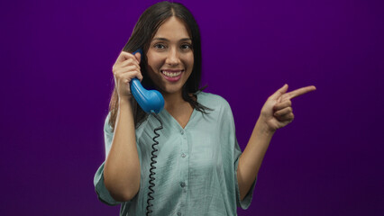 Brunette hispanic woman smiling holding blue handset to ear and pointing finger in studio with...