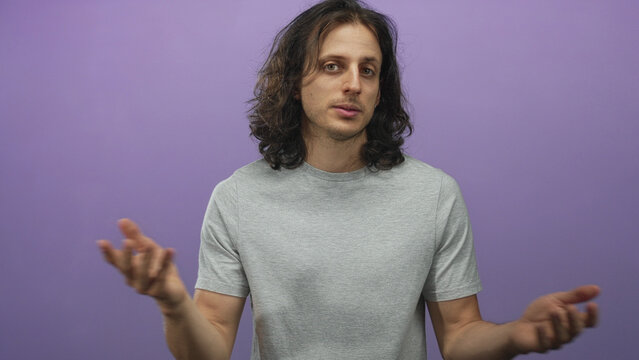 Man with long hair gesturing in purple studio, wearing grey t shirt and small earring, facing camera and speaking; calm reflection.