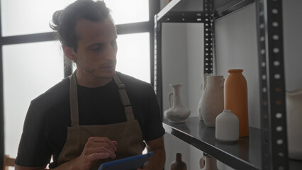 Man touching tablet while checking handmade ceramic vases on metal shelf in studio; craftsmanship steady concentration.