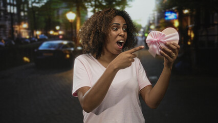 Woman points finger to heart shaped gift box on city street while holding it with bare hands and...