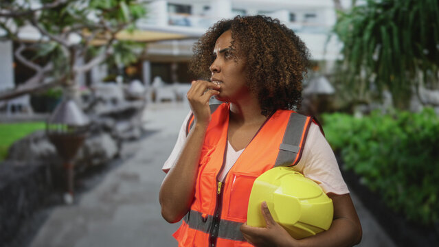 Woman engineer wearing orange safety vest bites nails while holding yellow hardhat on city street near building; anxiety uncertainty.