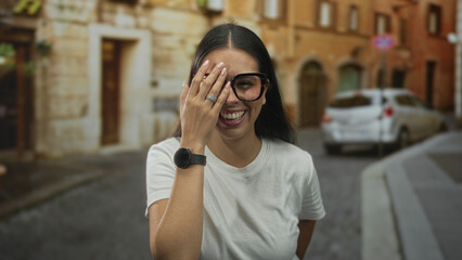 Fototapeta premium Woman smiling with hand covering one eye, wearing glasses and a white tee on a cobblestone street with parked car; joy carefree.