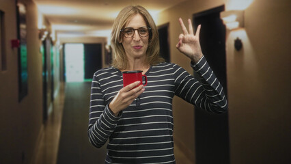 Woman holding red mug with right hand and making ok sign with left hand in building hallway; cozy...