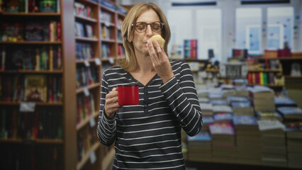 Woman holding a red mug and smelling a muffin with eyes closed, standing among bookshelves and stacked books inside a building  contentment. © Krakenimages.com