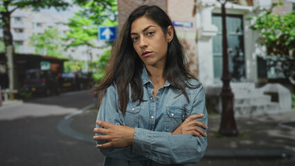 Fototapeta premium Woman arms crossed on city street in denim shirt, hands visible with ring and stern gaze; pensive resilience.