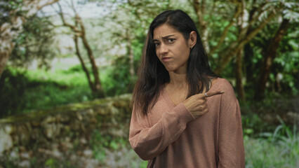 Young woman pointing finger in park beside a mossy stone wall, smiling and showing exposed index finger and v neck sweater while standing outdoors; serenity.