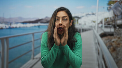 Woman in green sweater cups hands to mouth on seaside street promenade by the water calling out...