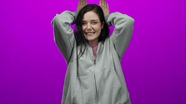 Woman with brunette hair playfully mimics animal ears against a vibrant pink background, wearing a casual gray hoodie, capturing youthful joy and creativity in a playful pose.
