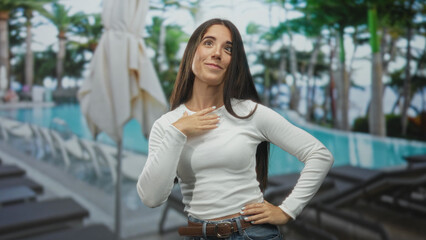 Young hispanic woman thumbs pointing to chest at resort pool wearing white long sleeve top and jeans by lounge chairs and umbrellas; confidence.