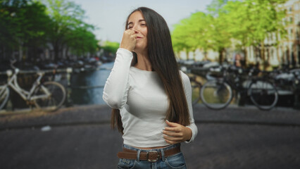 Young woman with hand to nose and expressive gesture on a street by a canal lined with bicycles; playful curiosity.