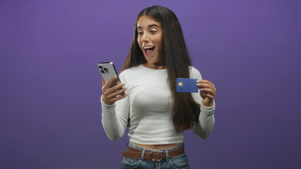 Young woman holds creditcard in one hand and smartphone in the other, hand visible, smiling in a studio; excitement.