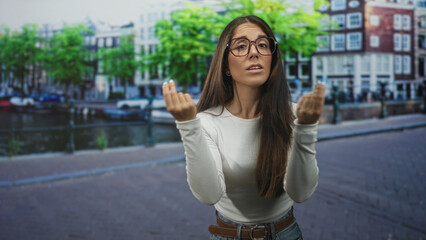 Woman with glasses beckons with hands on a canal street beside boats and railing, wearing white top and jeans with belt; pleading request.