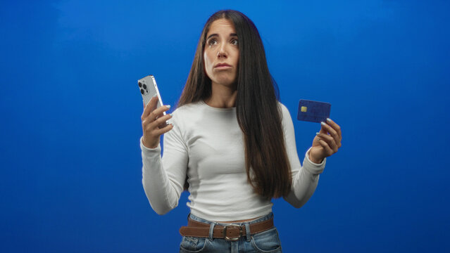 Woman holding smartphone and creditcard, shrugs and lifts hands while making a puzzled face against blue studio backdrop; payment uncertainty. - Powered by Adobe