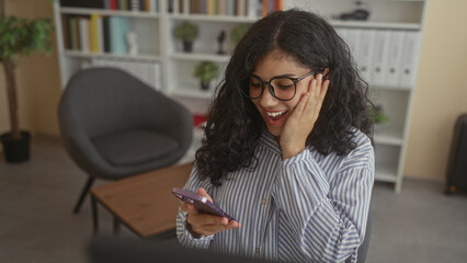 Young woman tapping smartphone with fingers and hand on cheek while smiling in office building near...