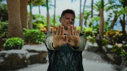 Man shielding face outdoors surrounded by lush greenery in sunny park setting showcasing defensive posture and visible discomfort in natural environment