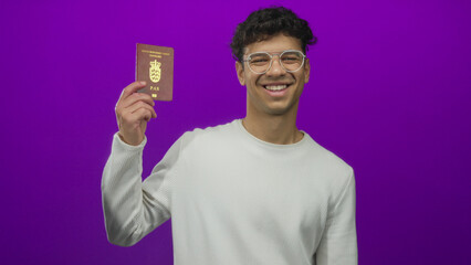 Young hispanic man raises hand holding a danish passport in purple studio; pride opportunity immigration.