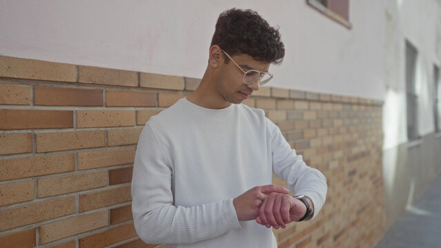 Man checking watch on street leaning against a sunlit brick wall while smiling; punctuality confidence.