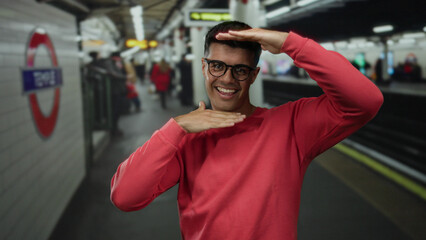 Obraz premium Young man wearing glasses making a framing gesture at a busy train station surrounded by commuters and bright lighting, capturing an urban scene.