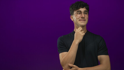 Young hispanic man wearing glasses and a black t shirt, hand on chin pensive pose in purple studio with slight smile and relaxed posture; thoughtful.