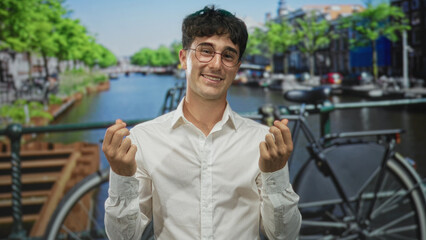 Man pinching fingers in money gesture on a street canal bridge beside a parked bicycle in amsterdam; playful optimism.