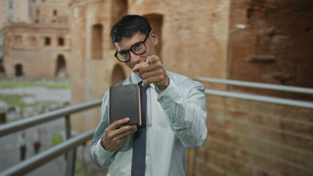 Man holds a leather book and beckons finger at ancient stone building ruins with a friendly smile; invitation. - Powered by Adobe