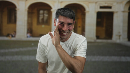 Man in white tshirt holds hand to mouth for whispering gesture in old town street with stone arches...