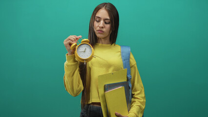 Young woman student holding yellow alarm clock and folders, presenting time with raised hand in studio; deadline urgency impatience.