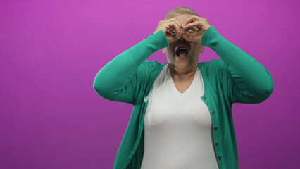 Middle aged caucasian woman making hand binoculars with hands over eyes in a purple studio wearing green cardigan and white tee; curiosity exploration.
