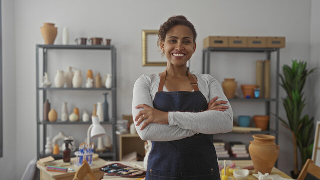 Woman standing with hands on hips wearing denim apron beside pottery shelves and vases in studio; creative pride. - Powered by Adobe