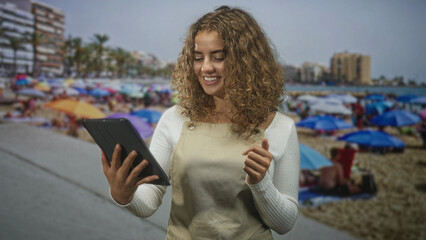 Woman holding tablet and gesturing with right hand, curly hair and apron, reading screen while smiling on street near crowded beach  contentment leisure. © Krakenimages.com