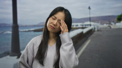 Woman outdoors on a seaside promenade looking pensive on a cloudy day with waves crashing, long hair blowing and lampposts lining the scenic beach promenade.
