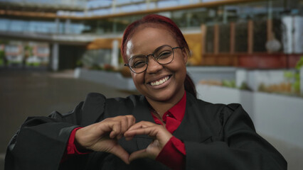 African american woman in black robe and red blouse smiles and forms heart with hands on street in daylight; love.